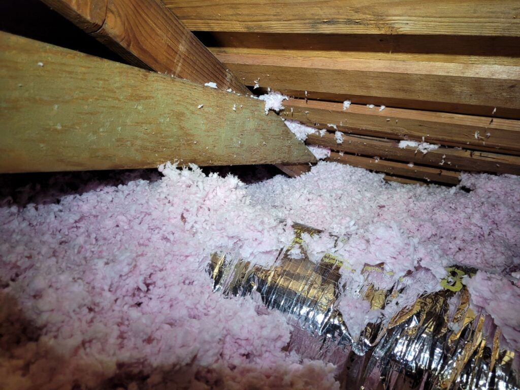Pink insulation in an attic, surrounding wooden beams and a silver duct.