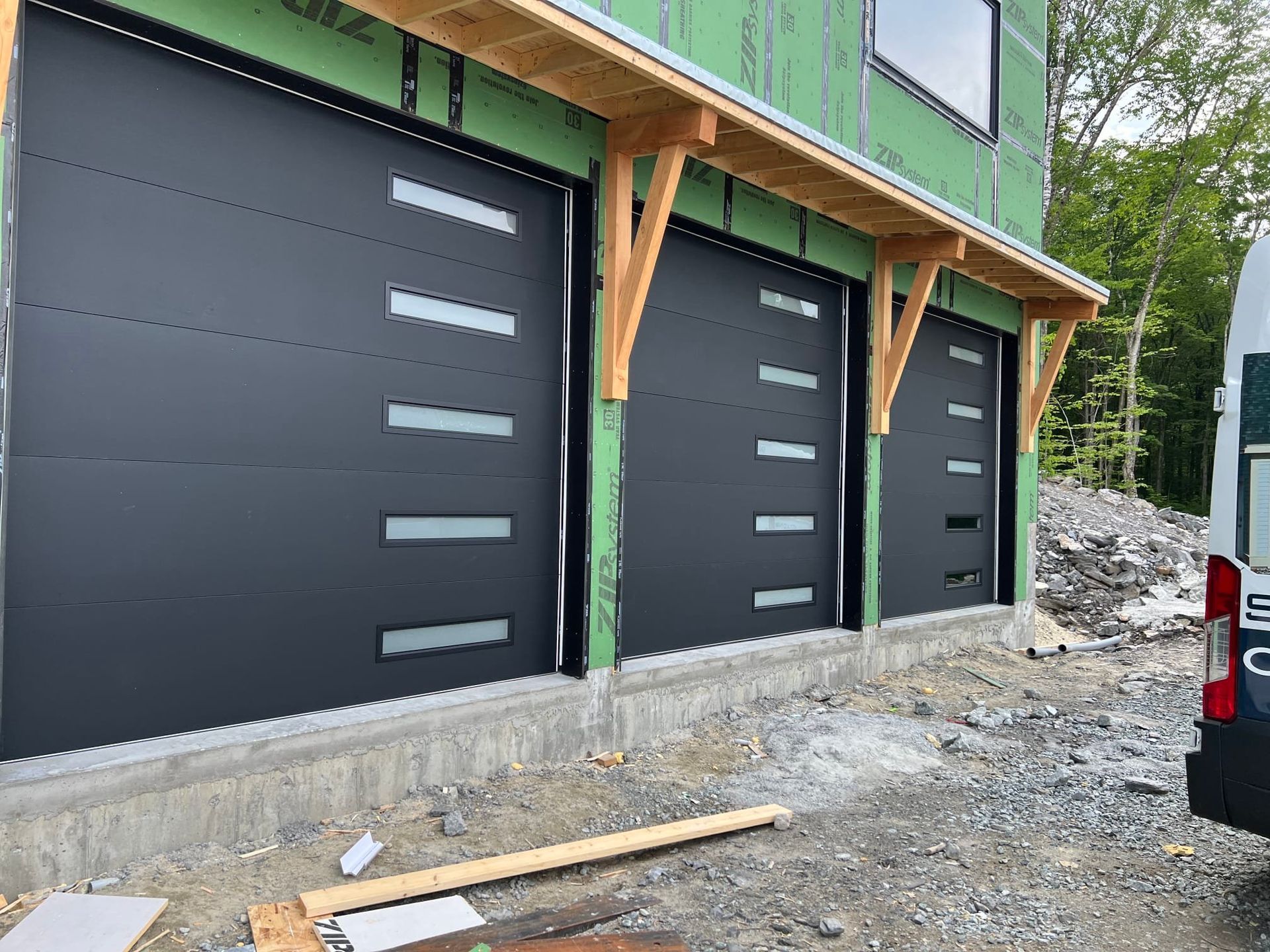 Three black garage doors on a green building under construction, with gravel and wooden supports in front.