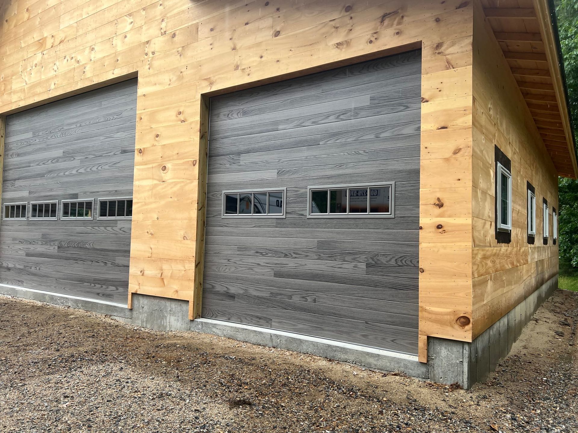 Two gray garage doors on a wood-sided building with gravel ground.