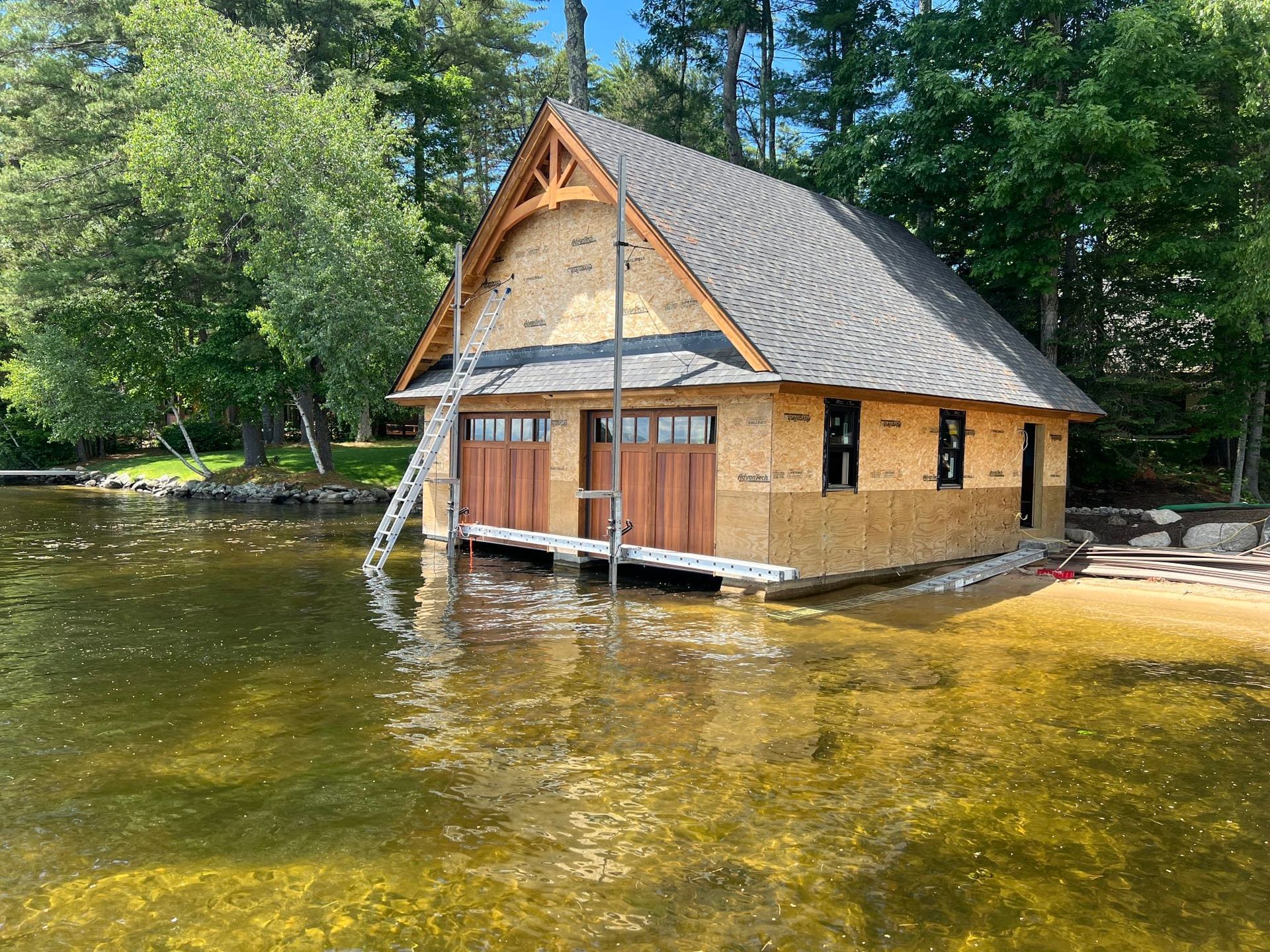 A small wooden boathouse with a gray roof sits partly in shallow water beside trees.