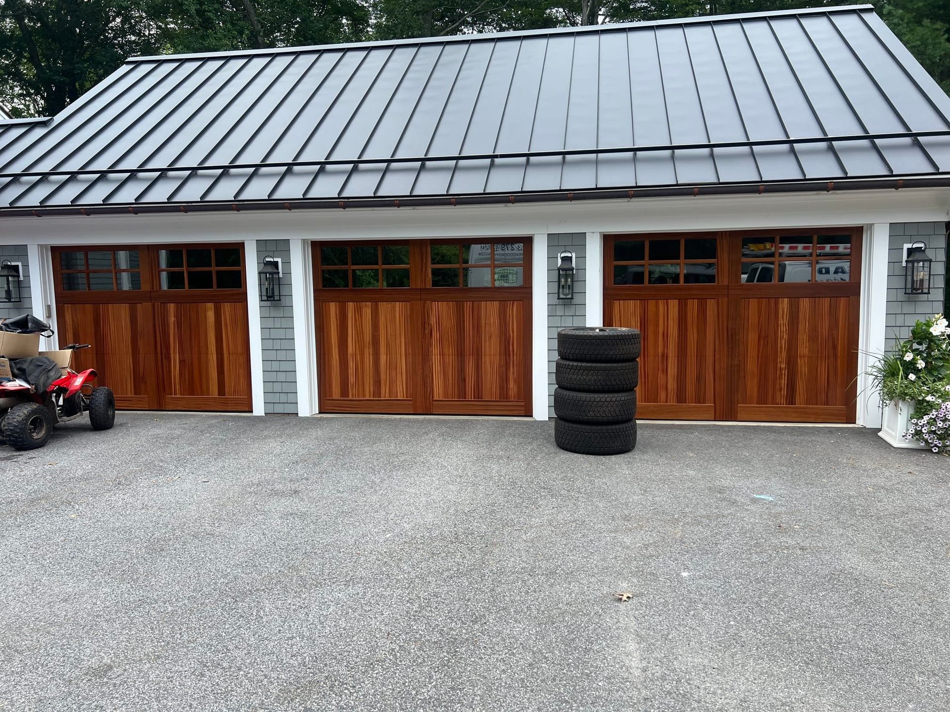 Three wooden garage doors on a gravel driveway with a gray metal roof above