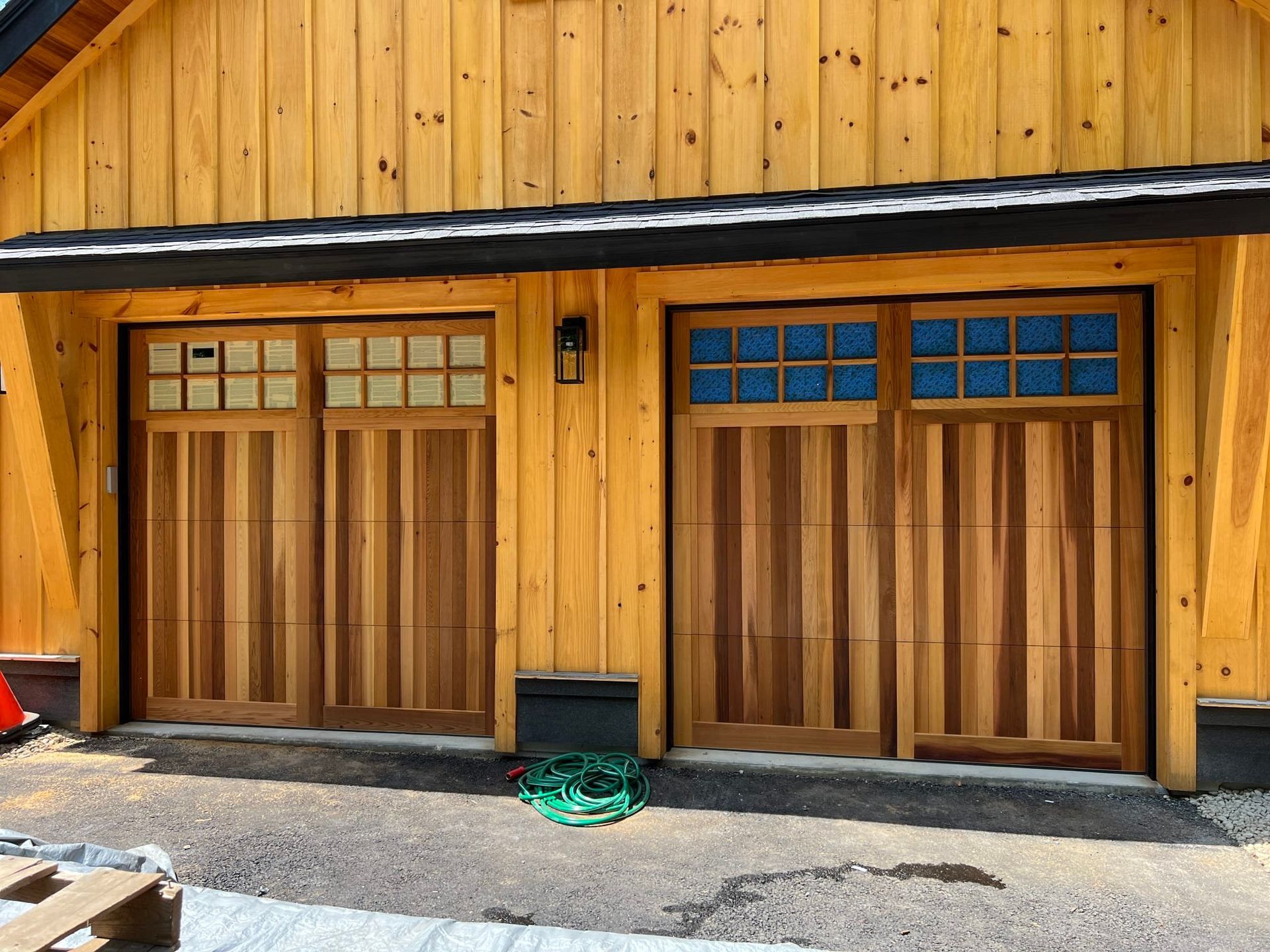 Two wooden garage doors on a yellow wood-sided building with blue window panes and a hose on the ground