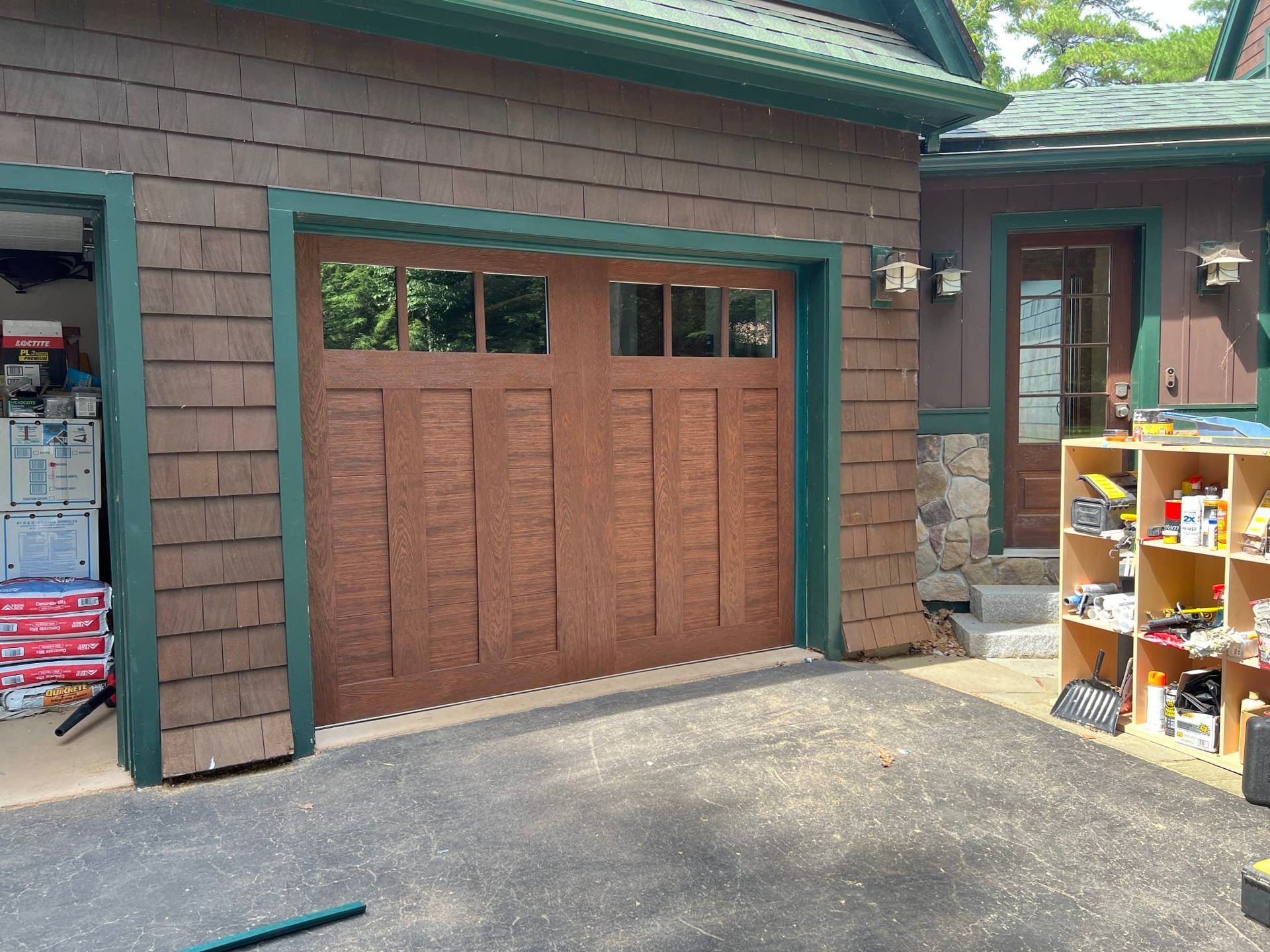Brown garage door on a cabin-style house with green trim, beside an open doorway and shelving outdoors.