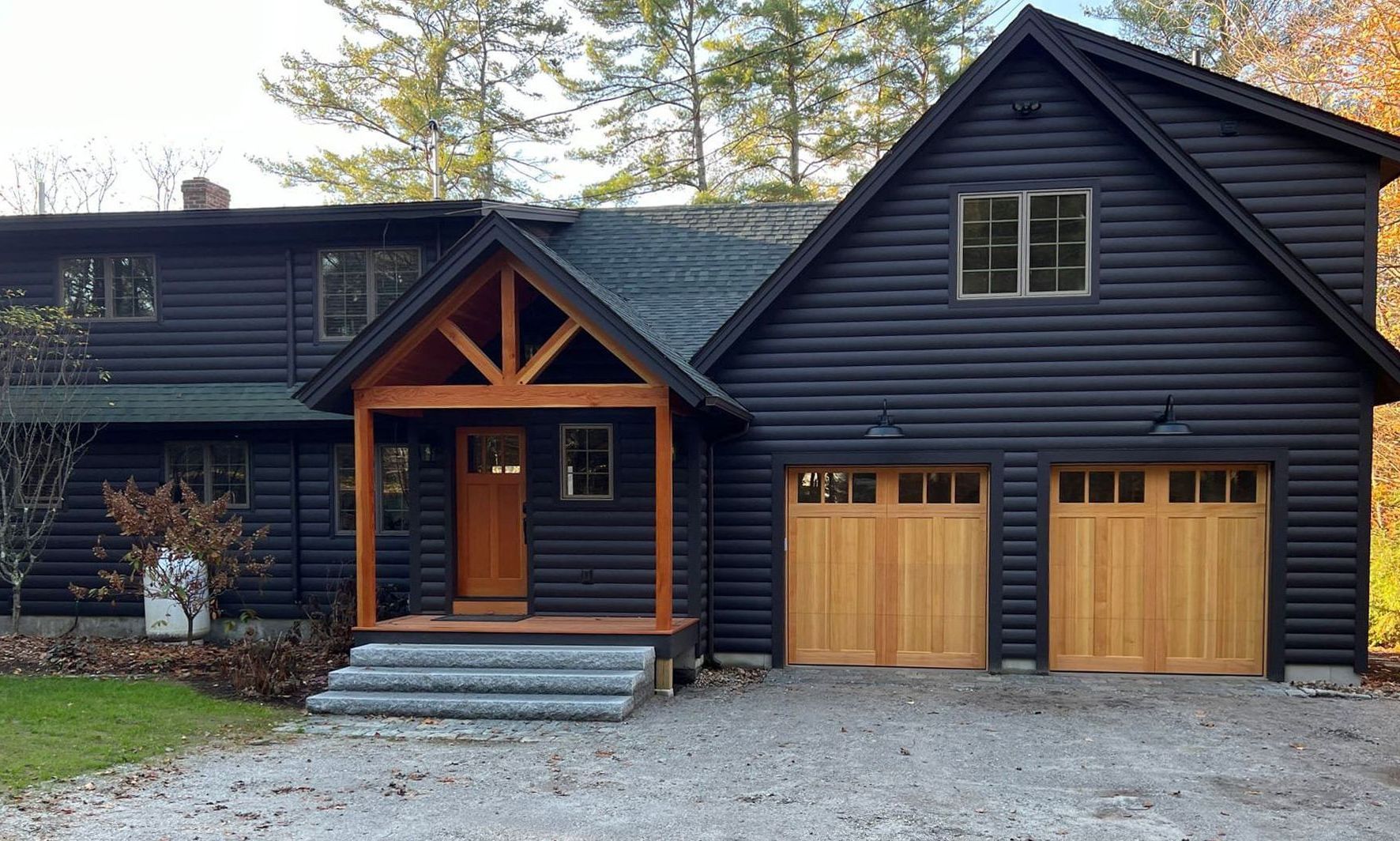 Black log cabin with light wood accents: front door, garage doors. Gravel driveway.