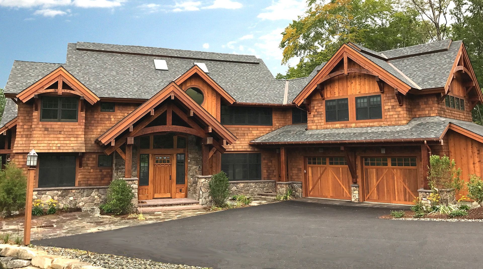 Brown wooden lodge-style house with a stone foundation, arched entry, and two-car garage on a paved driveway.