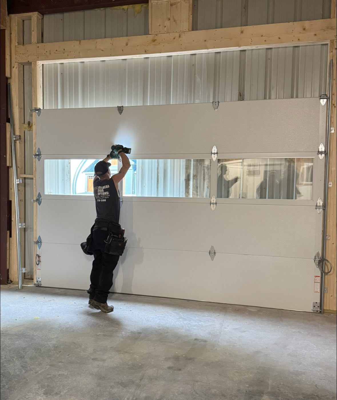 A construction worker installs a white garage door with a window panel in a warehouse.