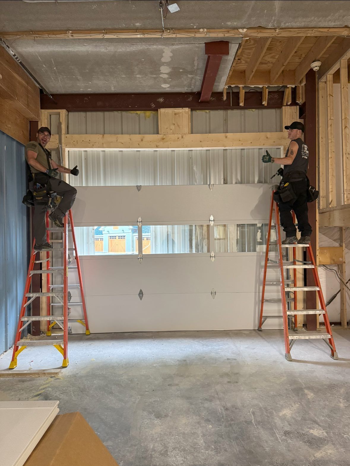 Two workers install drywall over a garage door opening, standing on ladders.