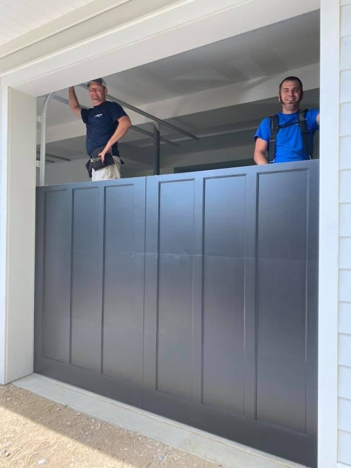 Two men installing a gray garage door. One holds a tool, the other looks at the camera. White trim around the door.