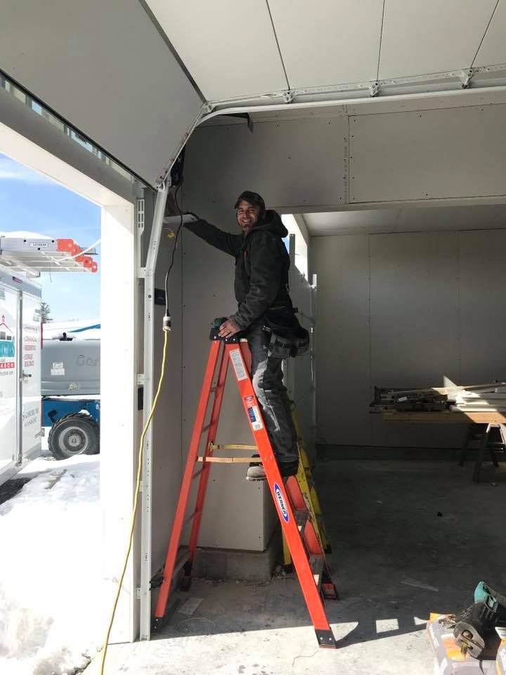 Man on orange ladder installing garage door; sunny, construction site.