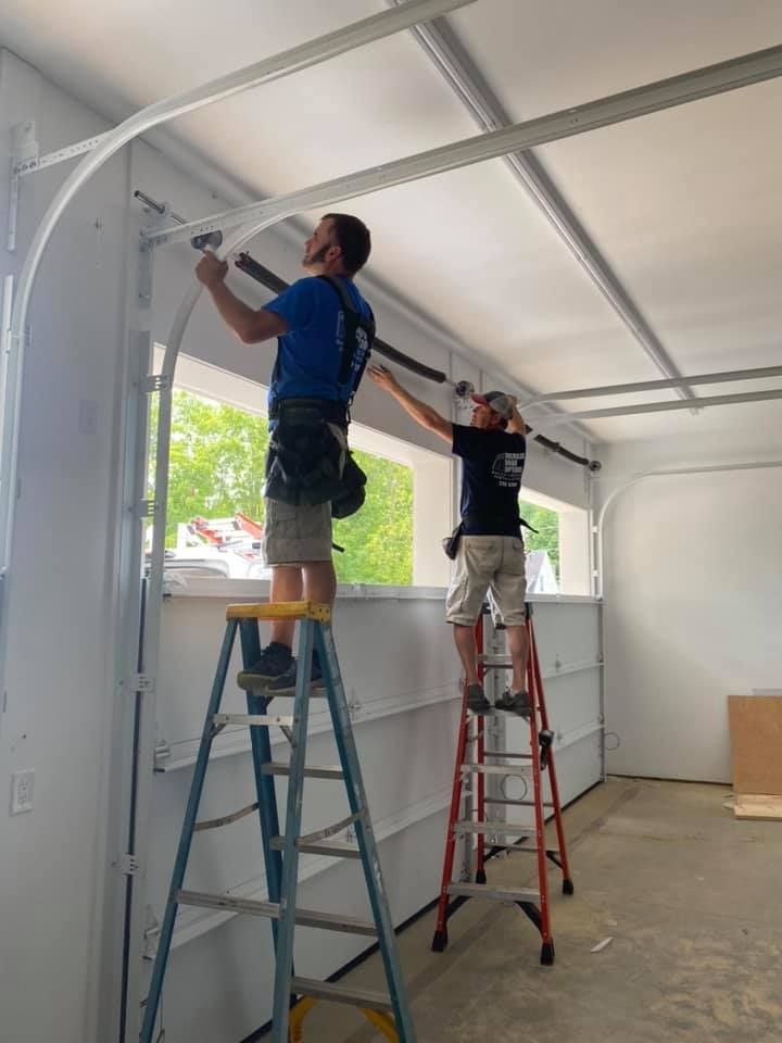 Two men installing a garage door, using ladders. They are wearing shorts and t-shirts in a white garage setting.