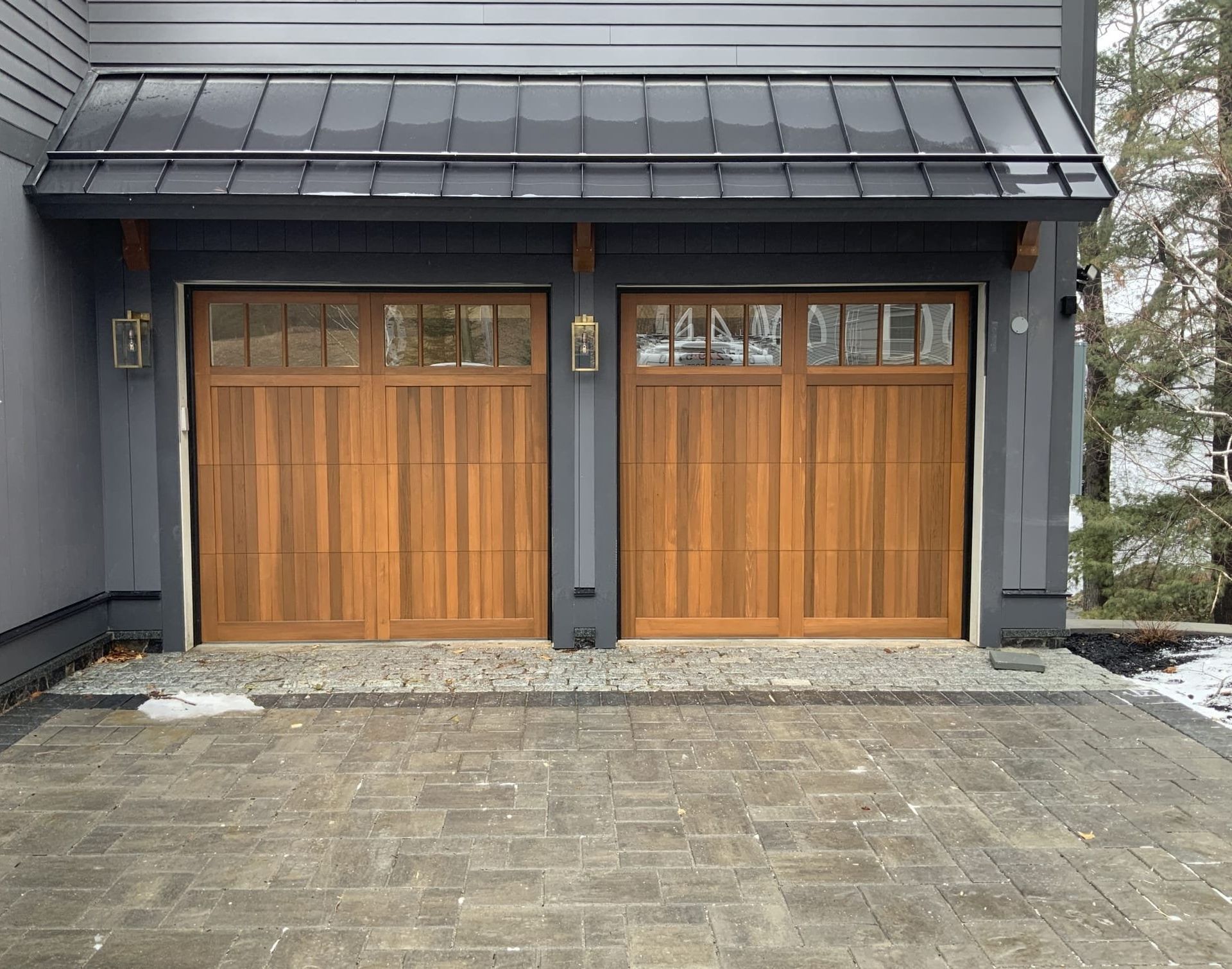 Two wood garage doors with windows, gray house exterior, black roof, and brick driveway.