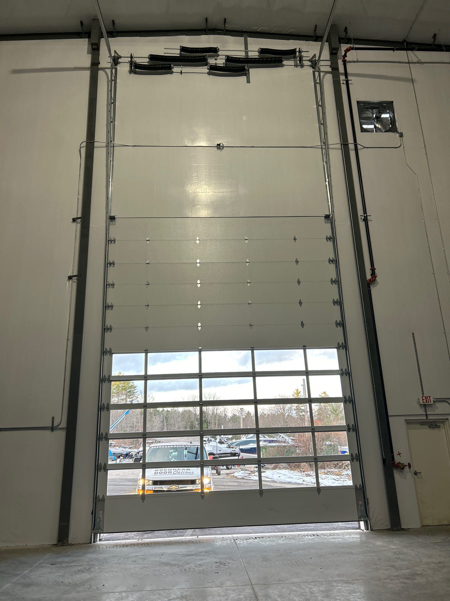 Open industrial garage door with clear window panes, view of parked cars outside.