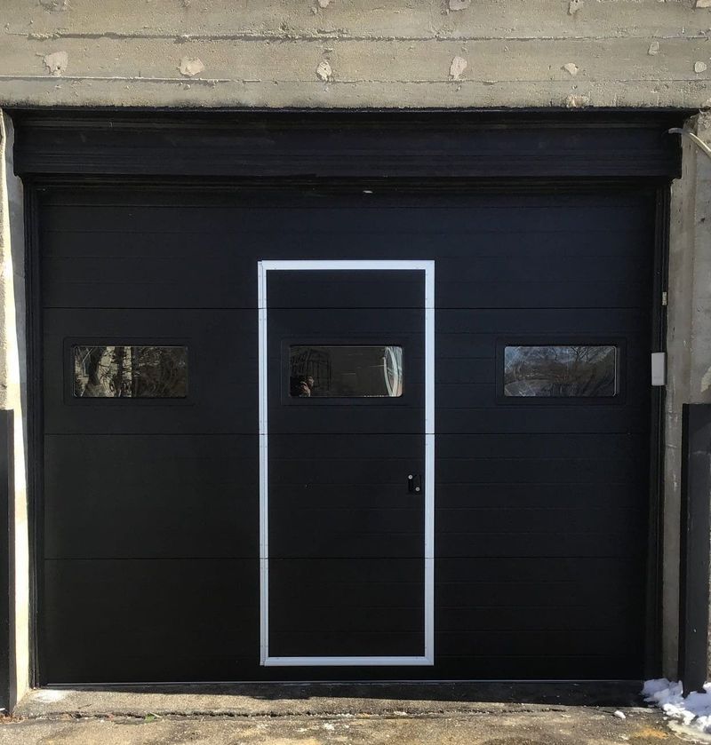 Black garage door with a centered walk-in door and windows; set in a concrete building.