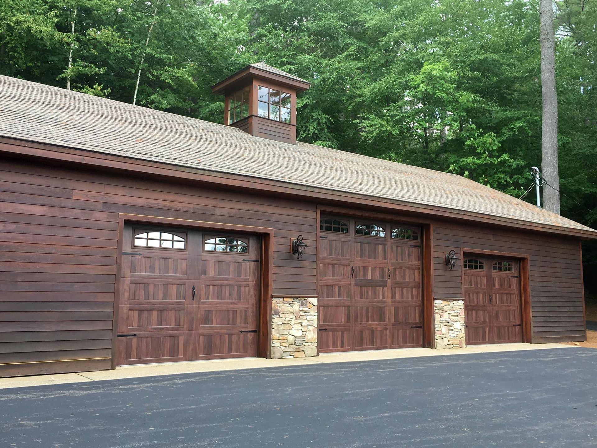 Brown wood garage with three doors, stone accents, and a cupola, set in a wooded area.
