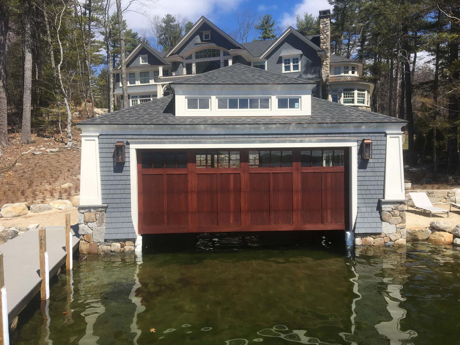 A large, gray boathouse with a red door sits on a lake, with a house on top.