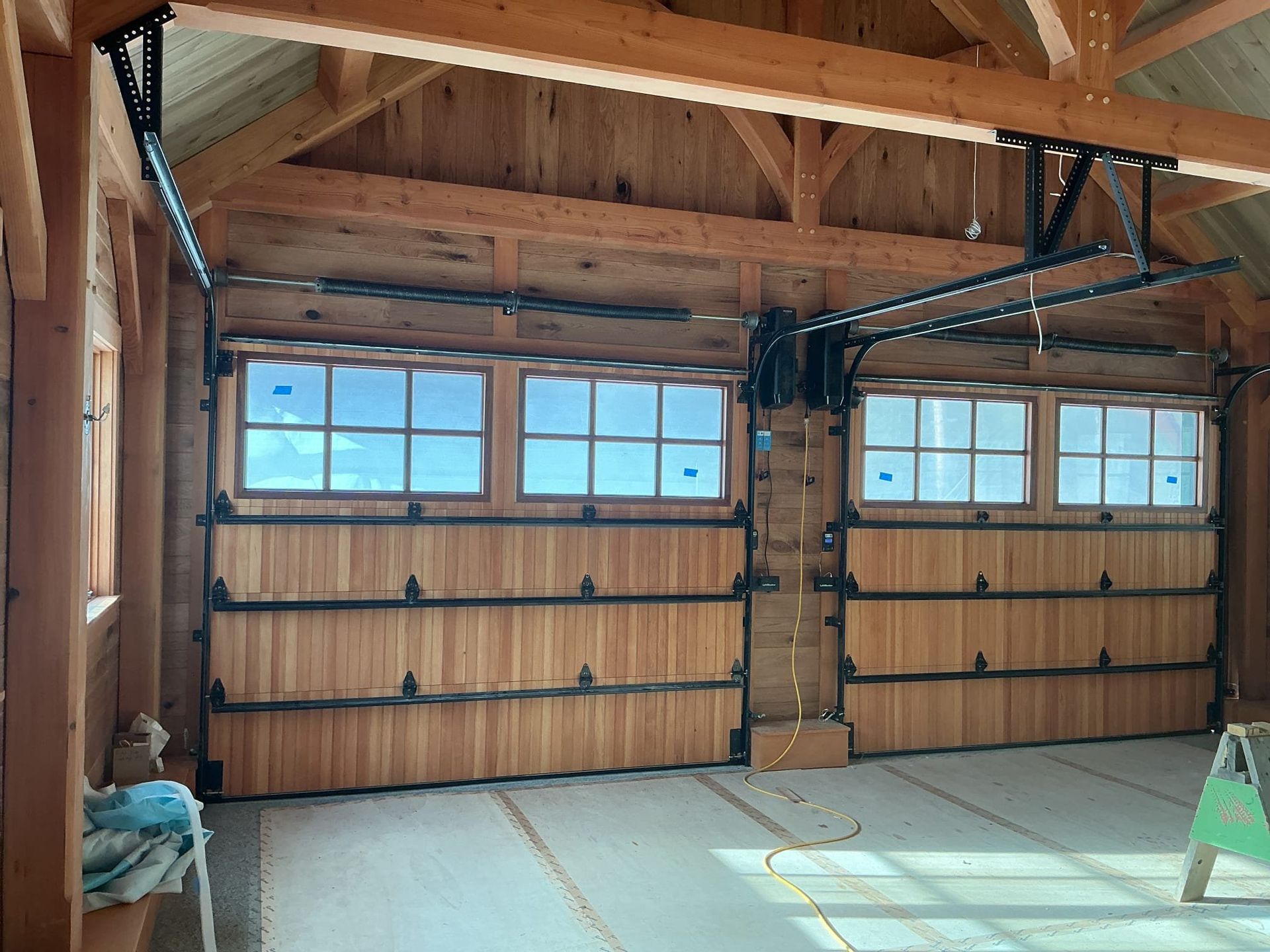 Two wooden garage doors with windows, mounted in a wooden-framed structure, open on a concrete floor.
