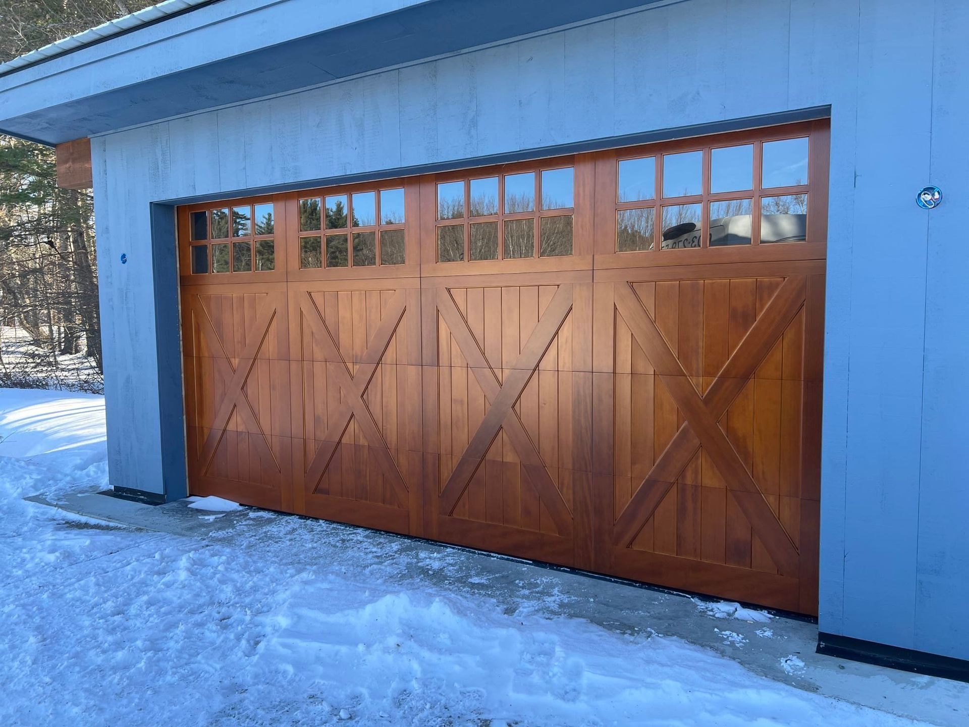 Wooden garage door with window panels, cross-shaped design, set in snow against a blue building.