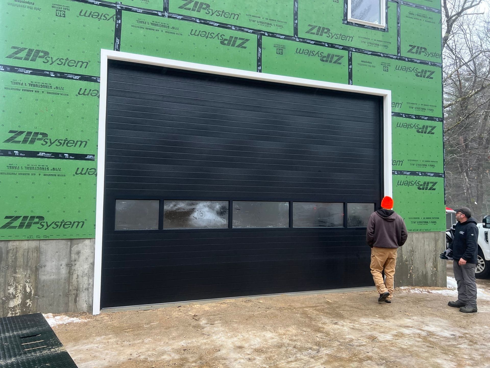 Black garage door with white trim, under construction. Two people standing in front of it.