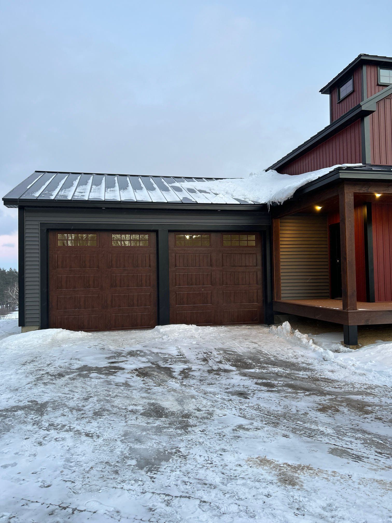 Two-car garage with brown doors, attached to a red house, snow on the ground and roof.