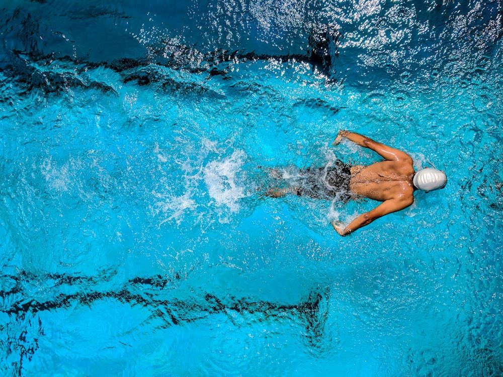A swimmer wearing a swim cap performs a butterfly stroke in a bright blue pool, viewed from directly above.