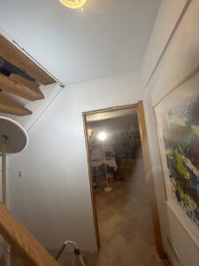A view looking down wooden stairs toward a doorway leading to a lit room, with a colorful abstract painting on the wall.