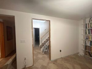 A room with off-white walls, a doorway leading to a staircase, light-colored tile floors, and a floor-to-ceiling bookshelf.