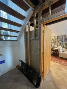 An unfinished wall frame next to a staircase and an open door revealing a room with a large bookshelf and seating area.