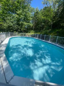 An oval-shaped swimming pool with blue water, surrounded by a white fence and lush green trees under a clear sky.