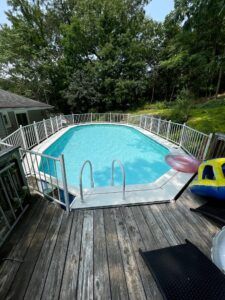 An above-ground pool with a white fence, surrounded by a wooden deck and trees.