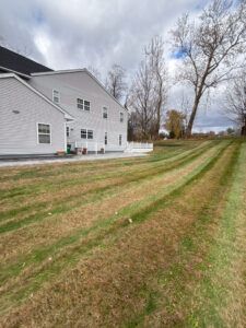 A multi-story beige house with a patio stands next to a lawn featuring mowed grass stripes leading toward bare trees.