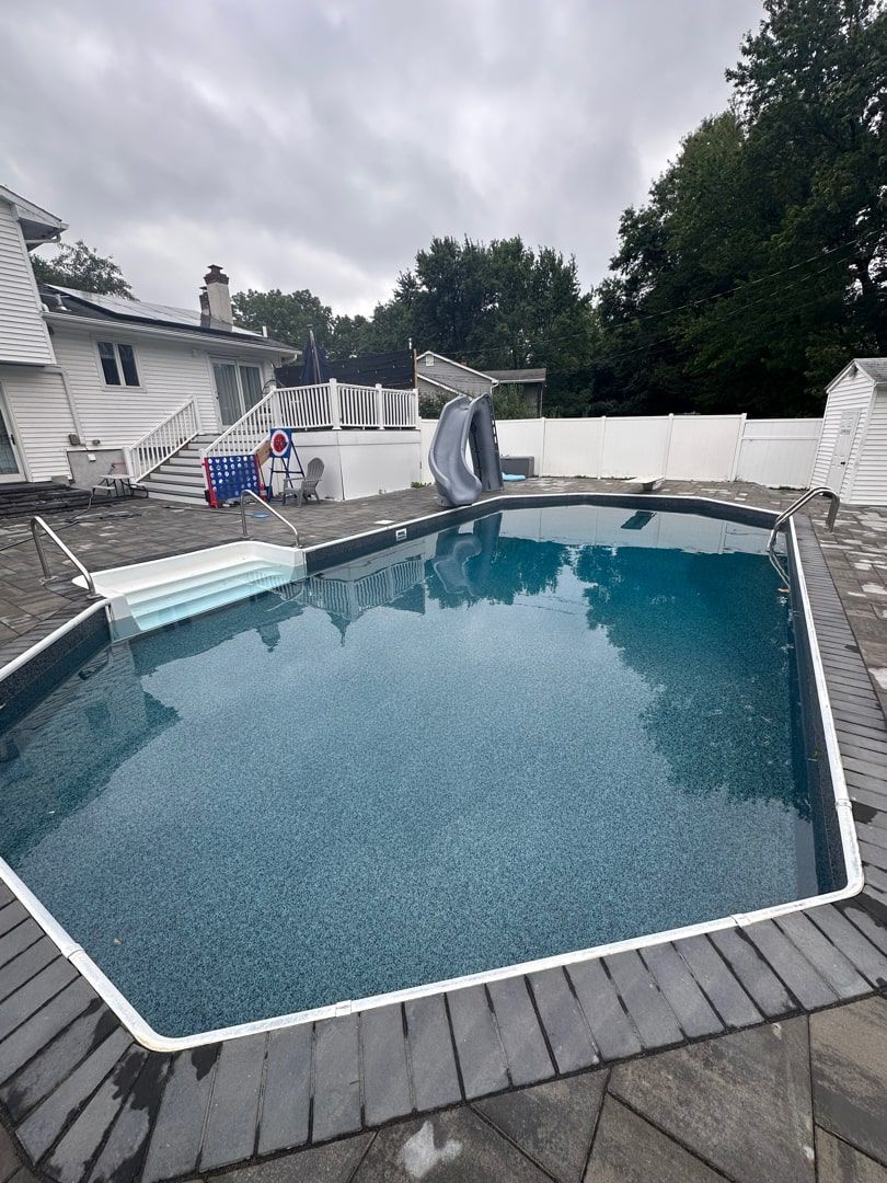 An outdoor swimming pool with dark blue water and gray stone coping, set in a backyard with a white fence and house.