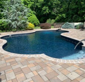 A blue, kidney-shaped swimming pool with a stone paver deck, surrounded by lush green trees and shrubs.