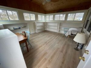 A sunroom with wood-paneled walls and ceiling, light wood flooring, white wraparound shelving, a small table, and a lamp.