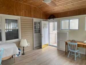 A bright sunroom with wood-paneled ceilings, light flooring, a small desk with a blue chair, and a table with a lamp.