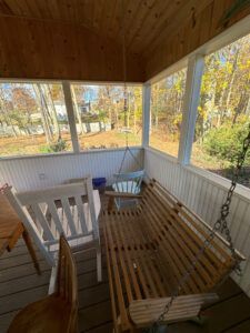 A screened-in porch with a wooden porch swing, a white wooden chair, and a view of trees through the screen.