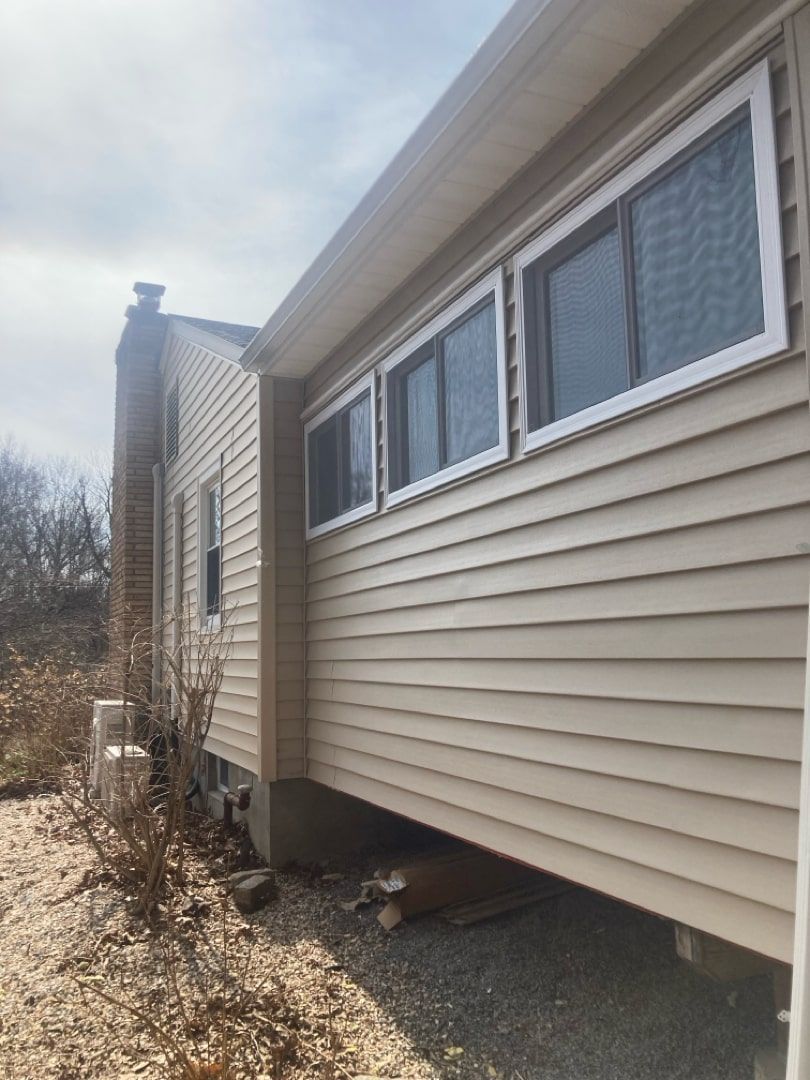 A side view of a beige-sided house with three windows above a crawl space and a gravel area.