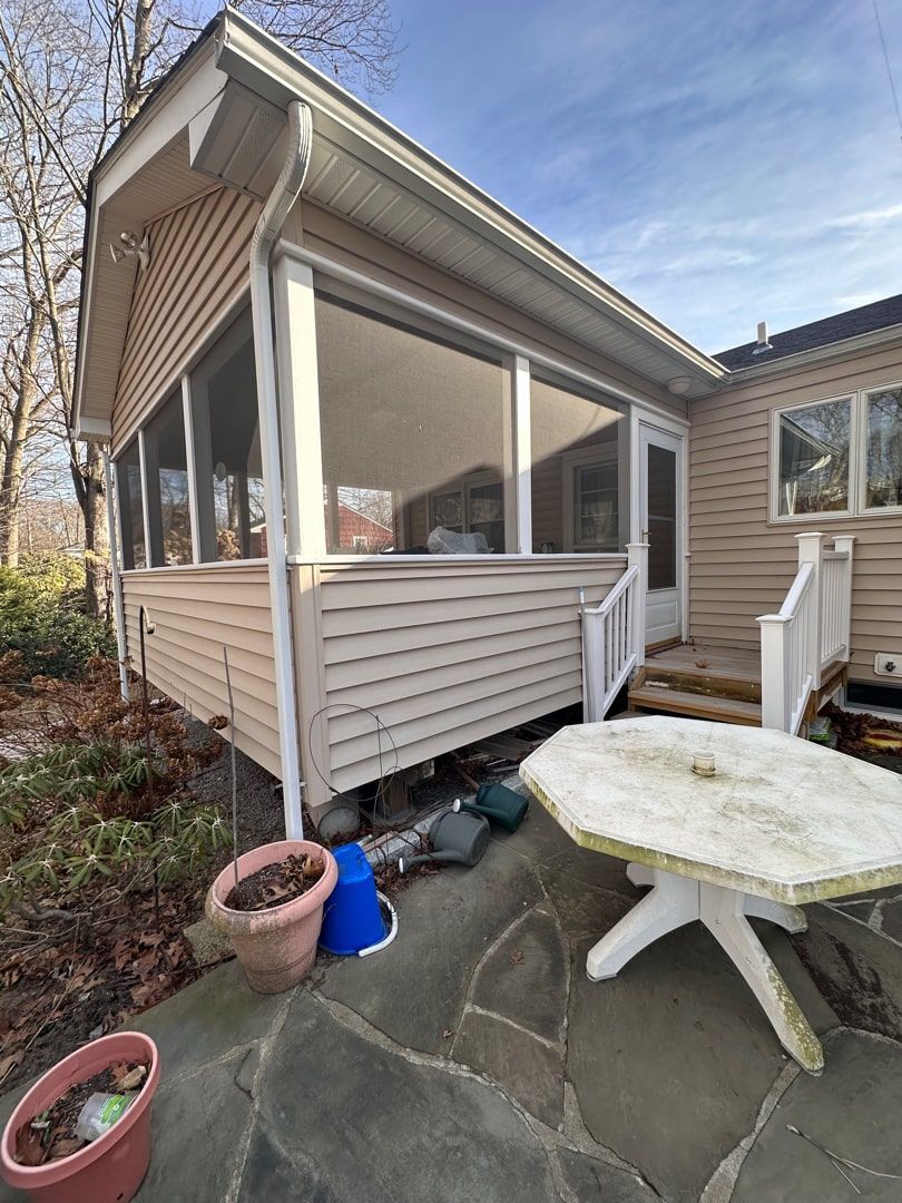 A screened-in porch with beige siding next to a stone patio featuring a white octagonal table and potted plants.