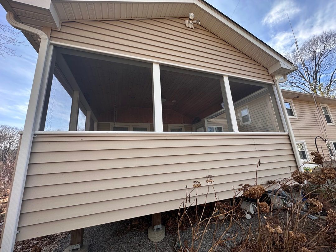 A tan, elevated screened-in porch with vinyl siding attached to a house against a blue sky with bare trees.