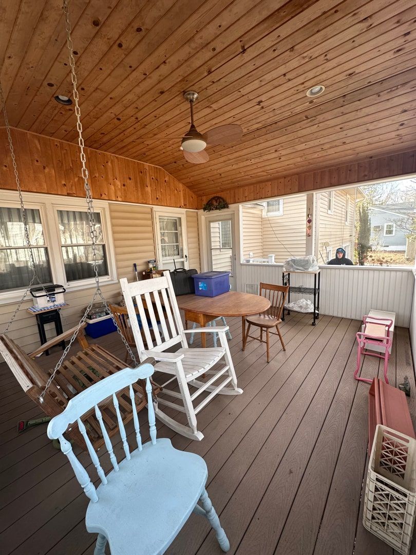 A covered outdoor porch with wood-paneled ceiling, a rocking chair, a light blue chair, a table, and other patio furniture.