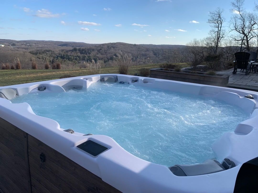 A steaming hot tub sits on a patio overlooking a vast, hilly landscape under a clear blue sky.