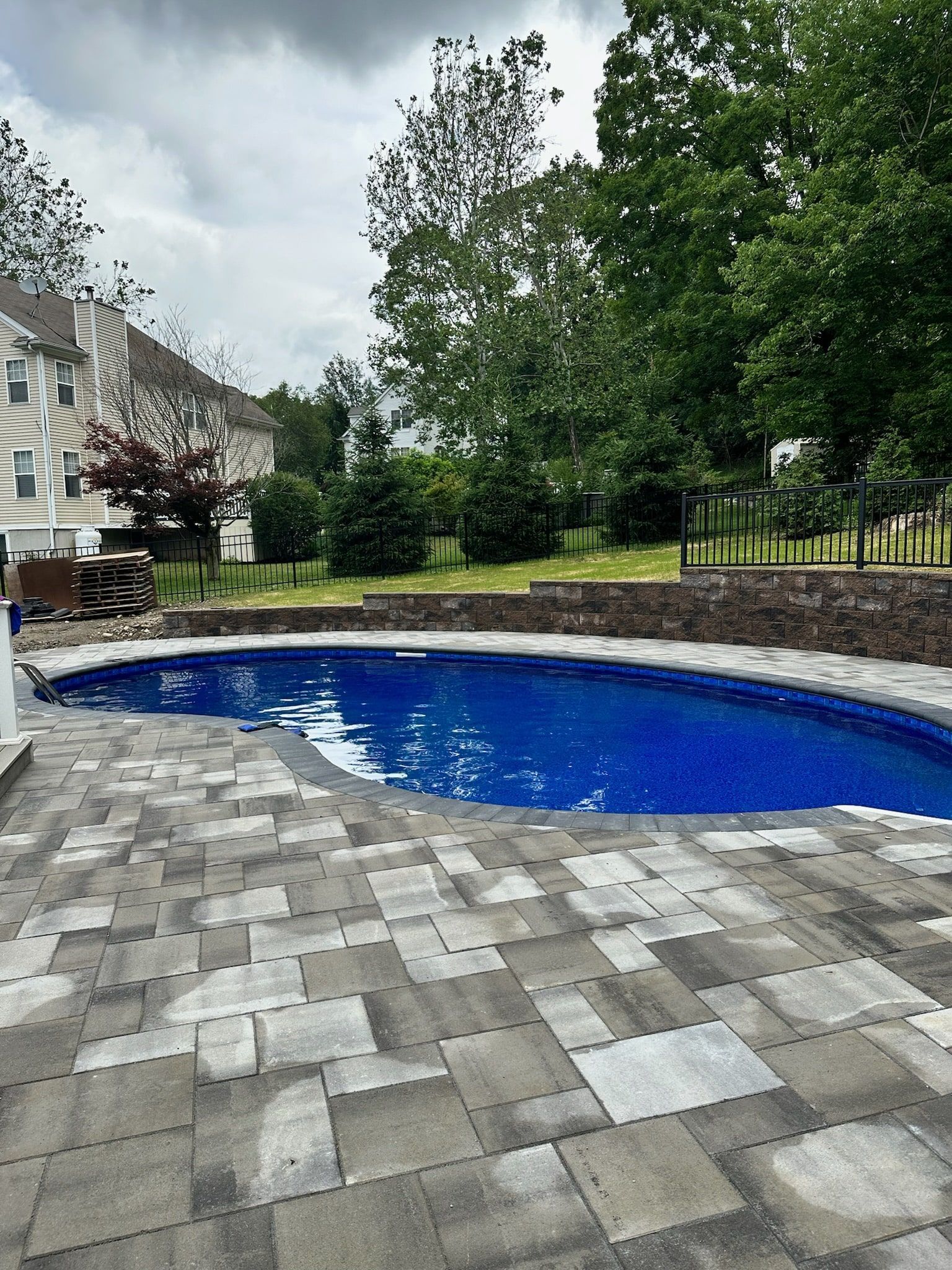 An oval swimming pool with bright blue water surrounded by a gray stone paver patio and a stone retaining wall.