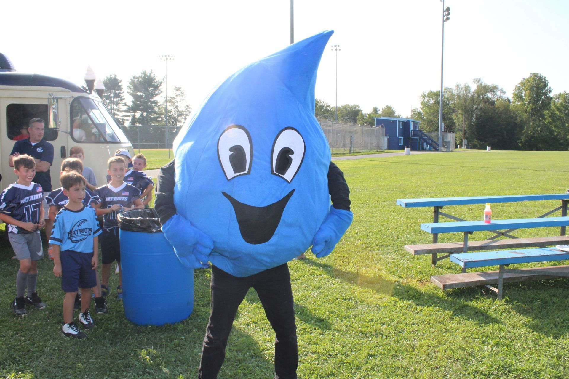 A person wearing a blue water droplet mascot costume standing in a field next to a group of children and a bench.