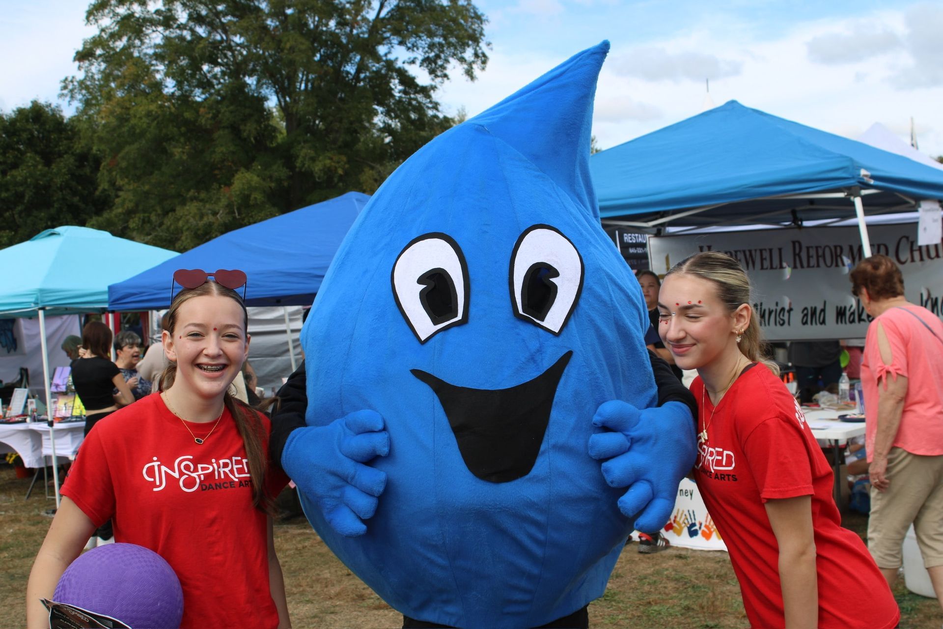 Two people in red shirts stand on either side of a large blue water droplet mascot at an outdoor festival.