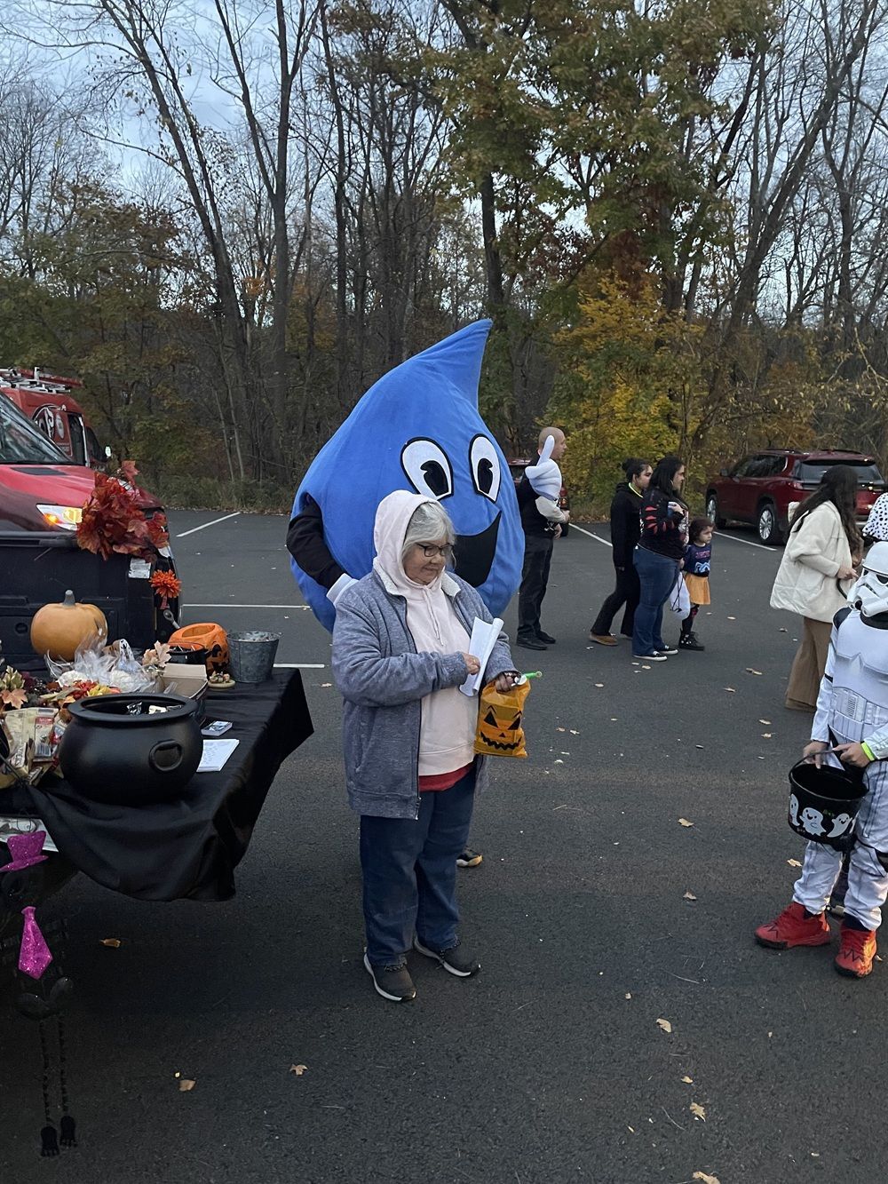 A person in a gray coat and a blue raindrop costume stand by a table with Halloween treats in a parking lot.