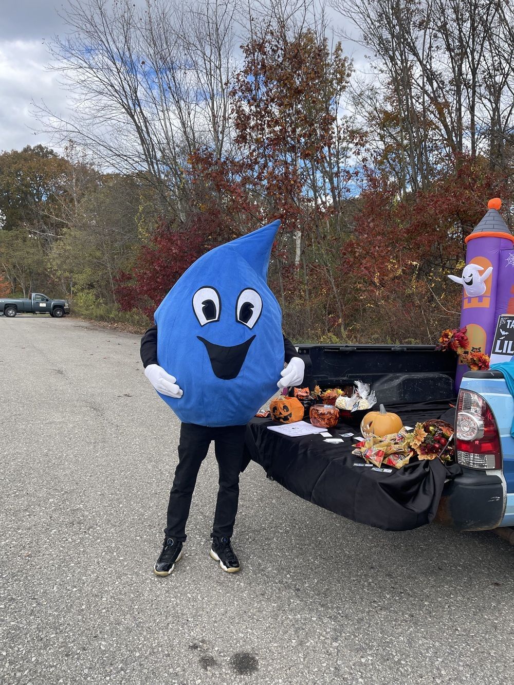 A person in a blue water drop mascot costume stands by the open tailgate of a truck decorated for Halloween.