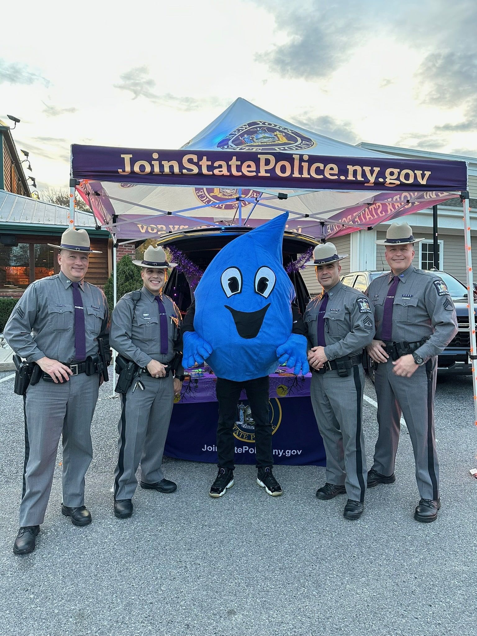 Four uniformed New York State Police officers stand with a blue mascot under a promotional tent at an outdoor event.