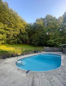 An oval-shaped swimming pool with blue water sits on a concrete patio in a backyard backed by a dense, green forest.