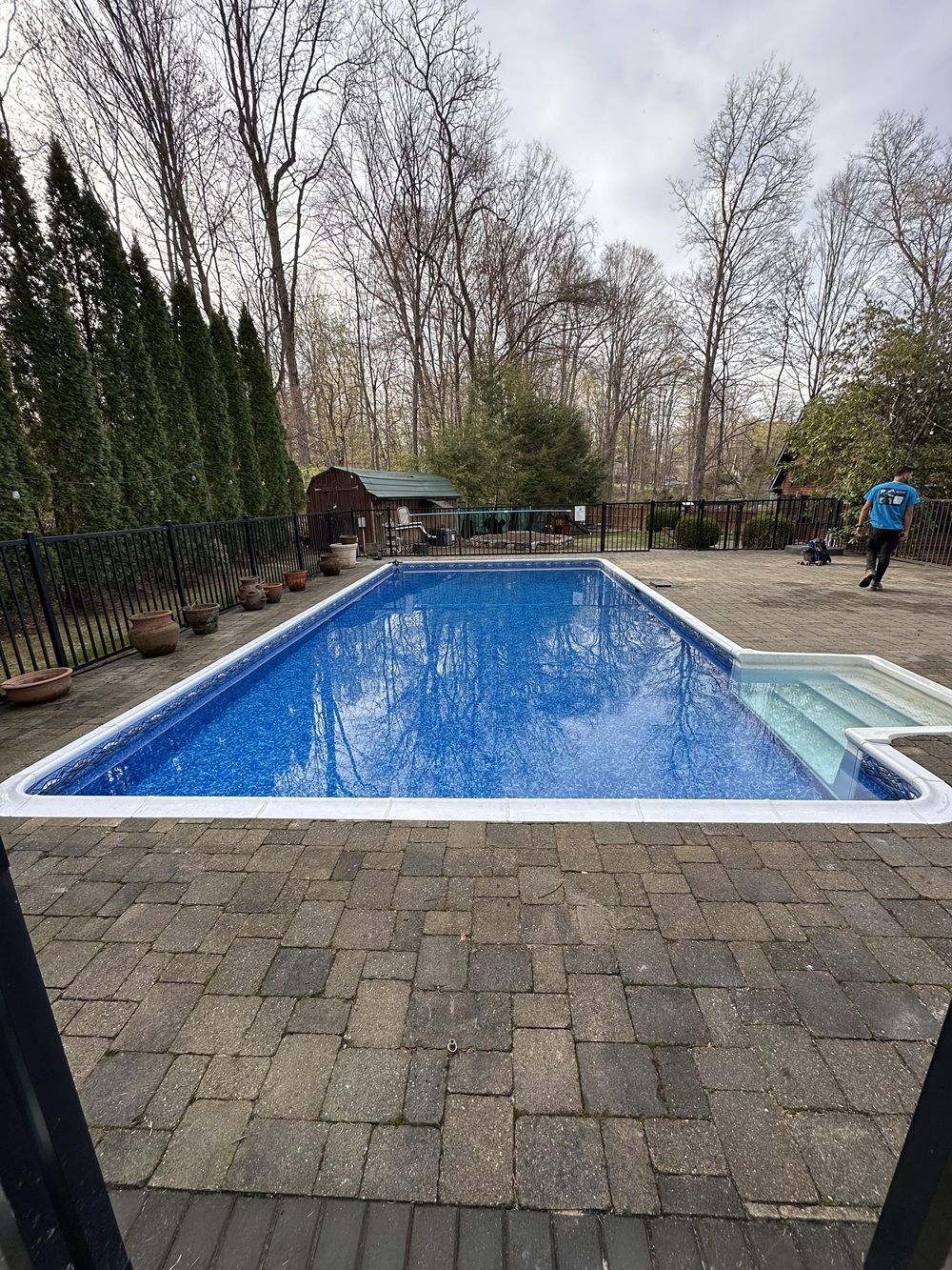 A rectangular, blue swimming pool with stairs, surrounded by a paver patio and woods, with a person walking in the yard.