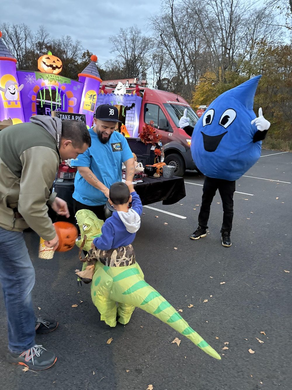 A child in an inflatable dinosaur costume receives treats from adults at a trunk-or-treat event with a blue mascot.