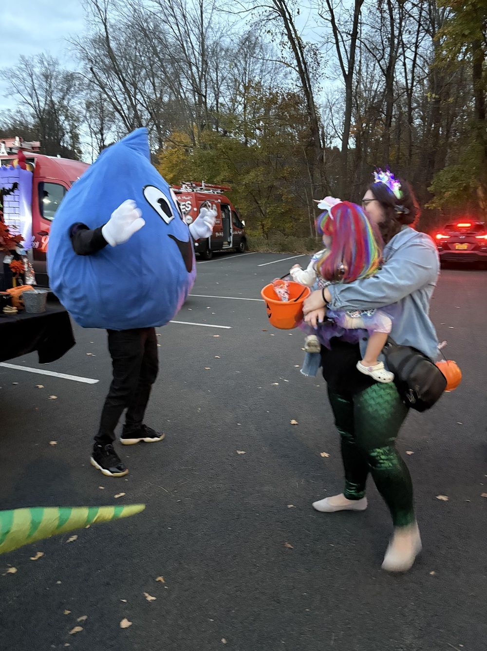A mascot in a blue raindrop costume interacts with an adult holding a child in a colorful unicorn costume at a parking lot.